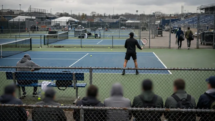 A generic tennis player practising alone on a tournament practice court while two generic players with racket bags walk away toward a distant court and fans watch through a chain-link fence, editorial graphic